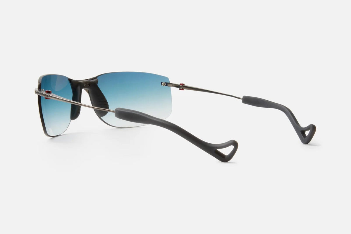 A pair of District Vision Miho Windstream Ti sunglasses by District Vision, featuring thin black titanium frames and blue gradient lenses, displayed from a rear side angle on a white background.