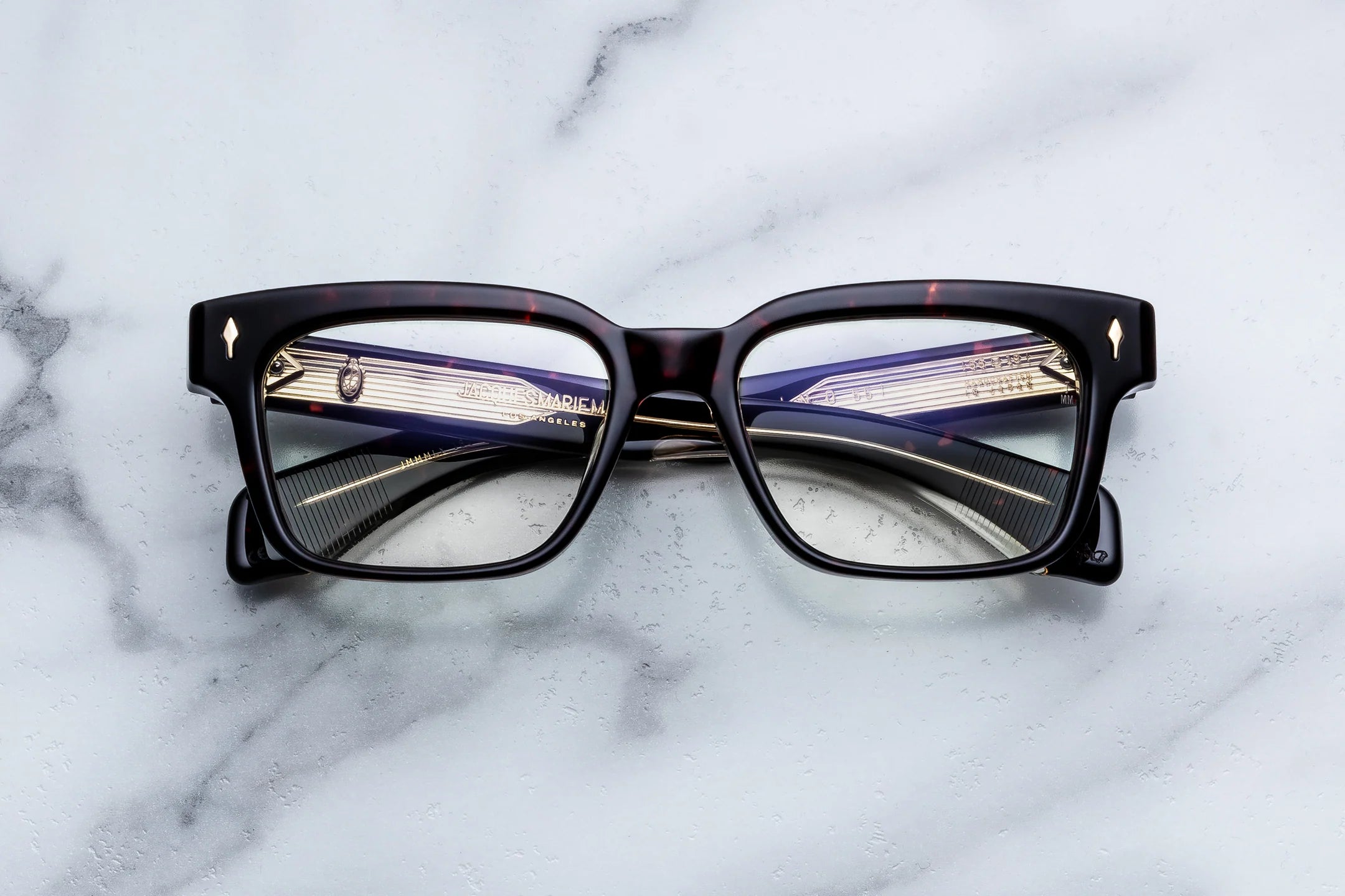 A pair of square-frame eyeglasses with thick black acetate rims and purple temple arms, resting on a white marble surface.