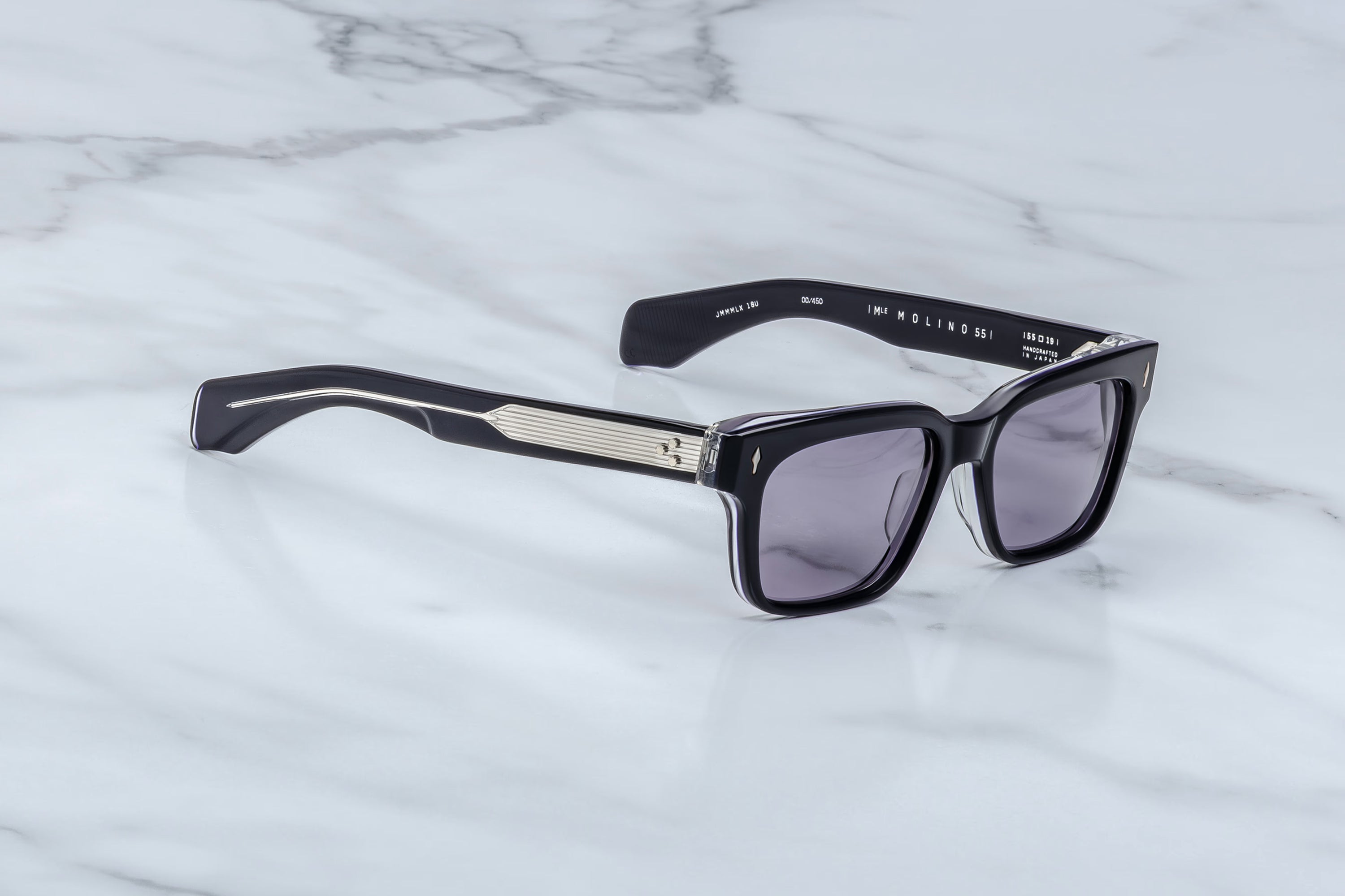 Black acetate sunglasses with dark gray lenses and a silver-tone geometric metal accent on the bridge, resting on a white marble surface.