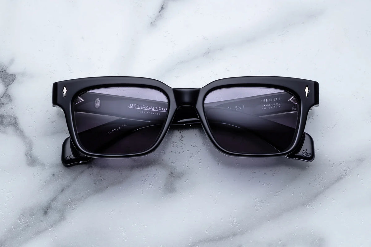 Jacques Marie Mage Molino 55 eyeglasses from Jacques Marie Mage feature thick black rectangular frames with subtle bevel detailing that enhances their classic silhouette. Resting on a white marble surface, the reflective lenses display faint digital text and icons, hinting at advanced heads-up display or smart glasses technology.