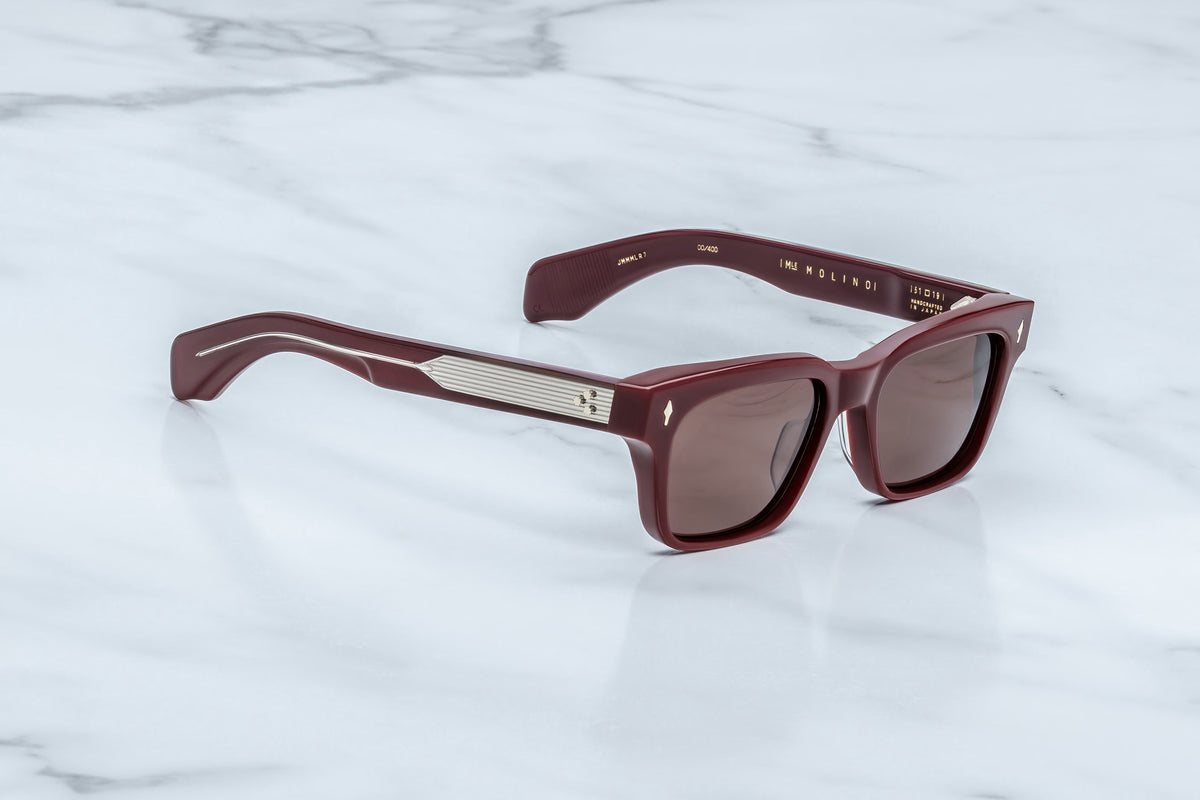 Jacques Marie Mage Molino sunglasses by Jacques Marie Mage, featuring maroon square frames with dark lenses and metallic accents on the arms—an homage to Carlo Mollino’s bold design aesthetic—resting on a white marble surface.