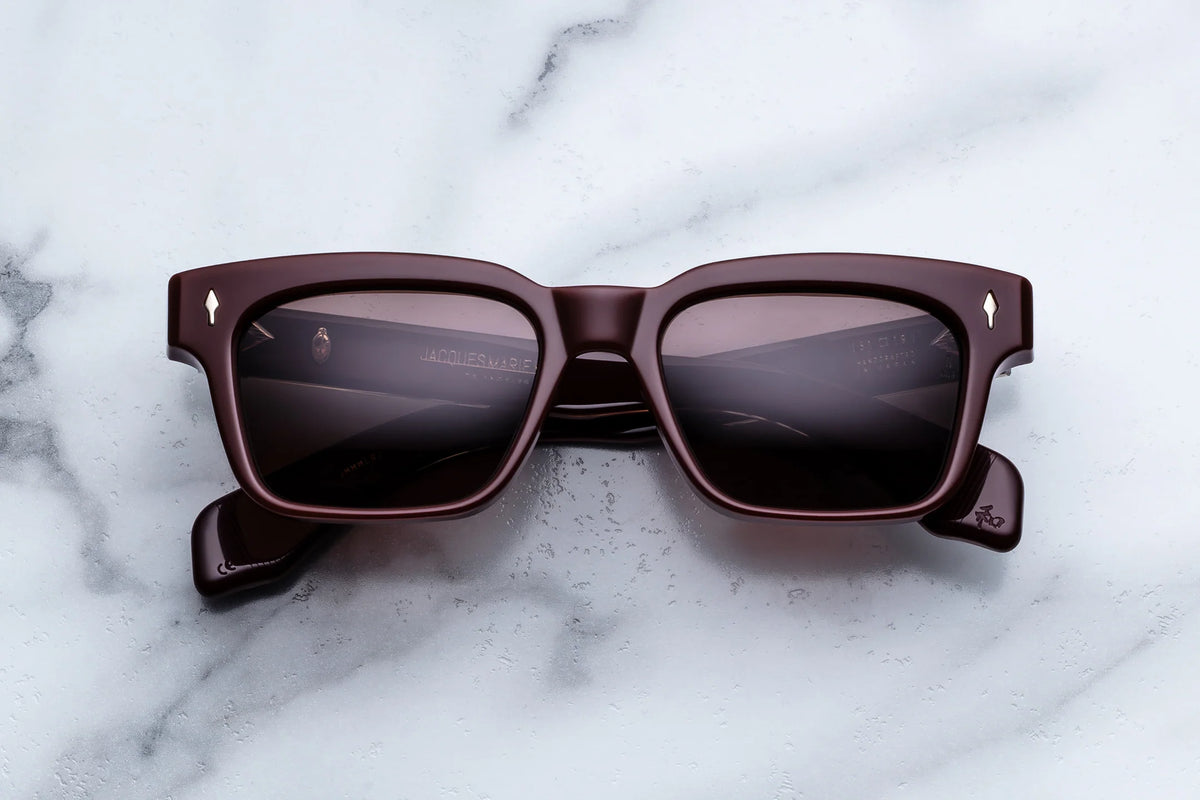 A pair of Jacques Marie Mage Molino dark brown rectangular sunglasses with dark lenses, inspired by Italian designer Carlo Mollino, rests on a light gray marble surface.