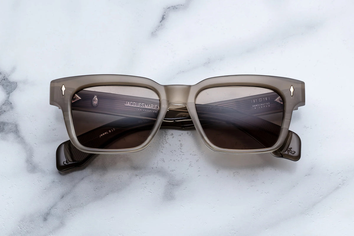 Jacques Marie Mage Molino sunglasses featuring thick, translucent brown square frames and dark lenses rest on a marble surface—a design that channels the refined sophistication characteristic of iconic Italian designer Carlo Mollino.