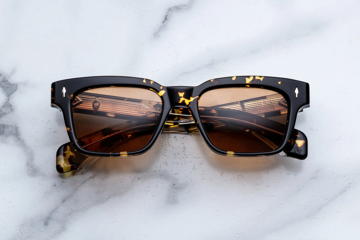 Jacques Marie Mage Molino tortoiseshell square sunglasses with brown lenses rest elegantly on a white marble surface. The arms and frame display a mottled pattern inspired by renowned Italian designer Carlo Mollino’s signature style, while the rectangular silhouette features lenses reflecting delicate gold lines, capturing the distinctive essence of Jacques Marie Mage craftsmanship.