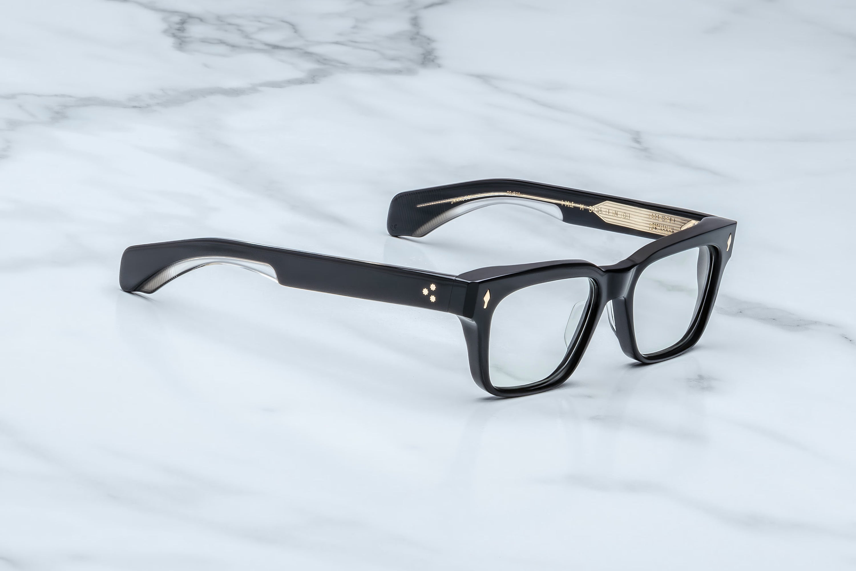 Black acetate square-frame eyeglasses with clear lenses and small gold geometric accents on the temples, resting on a white marble surface.