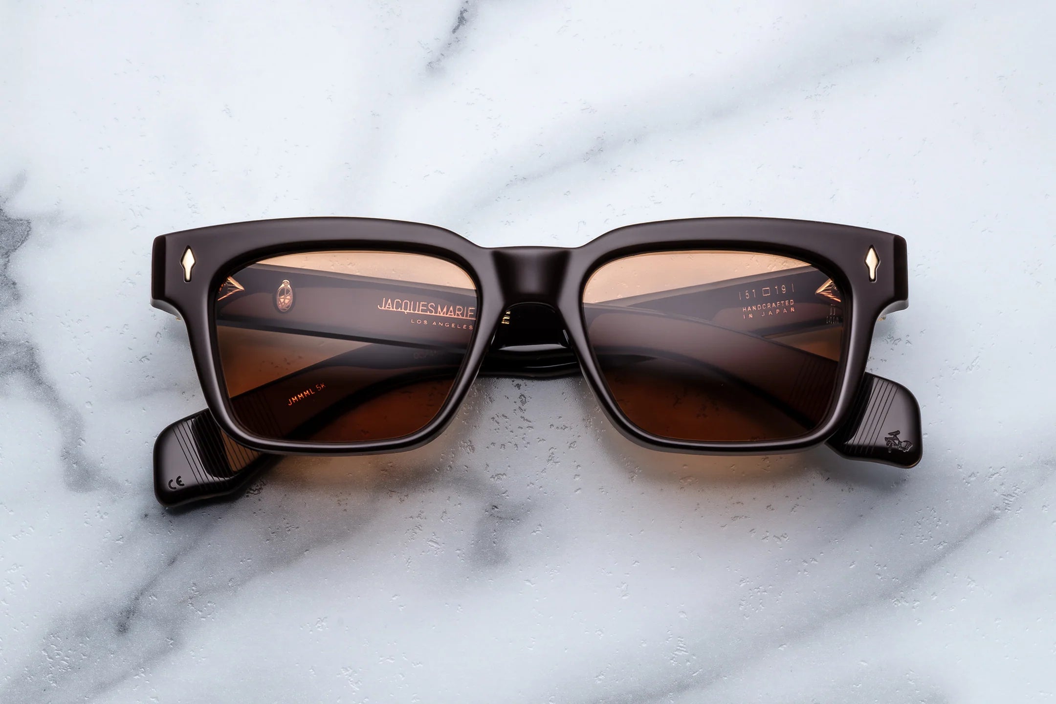 Jacques Marie Mage Molino rectangular sunglasses in dark brown, featuring brown lenses and subtle gold accents near the hinges, are displayed on a white marble surface. These frames reflect the refined elegance of Italian designer eyewear inspired by Carlo Mollino’s sophisticated aesthetic.