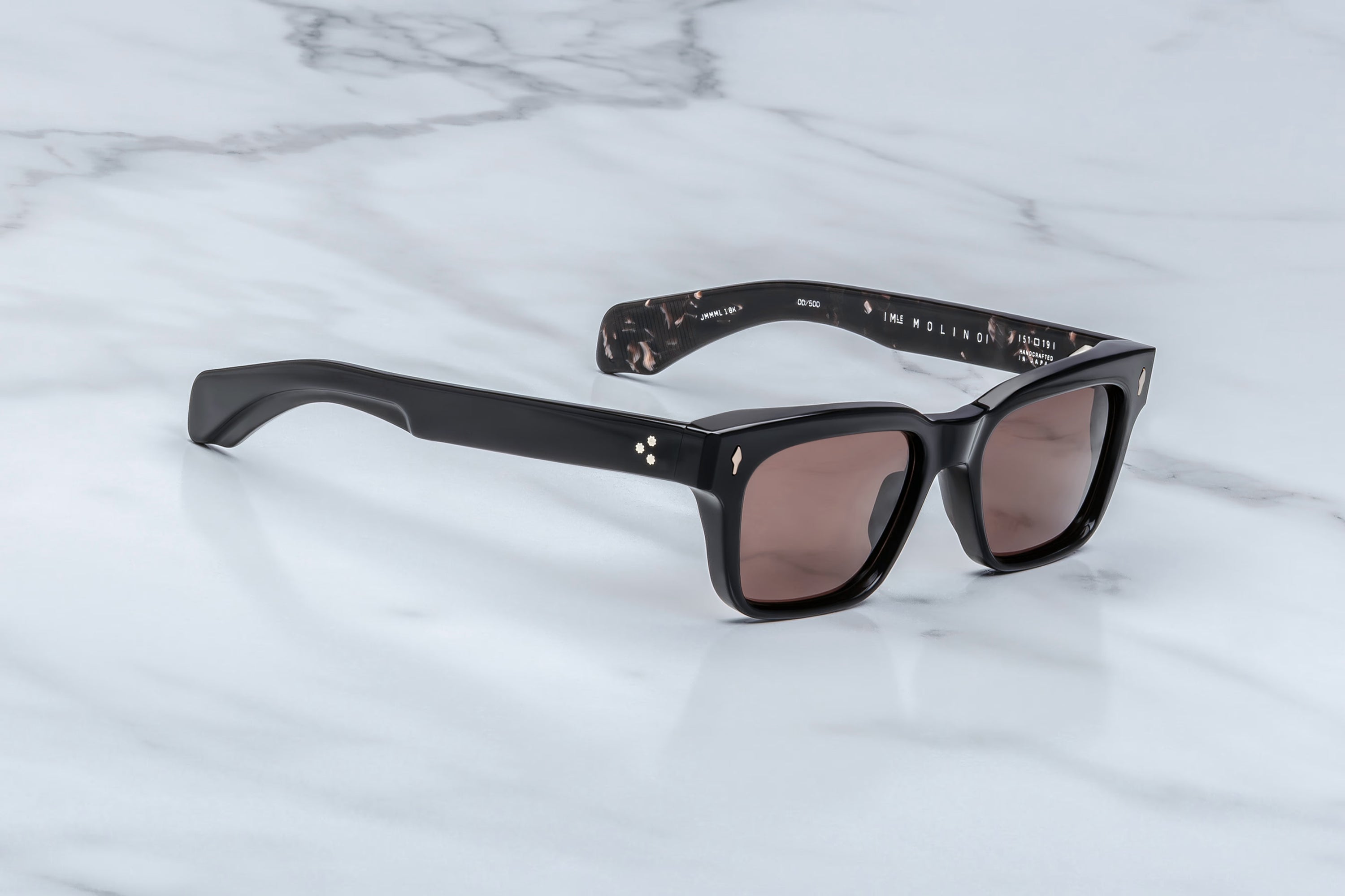 Jacques Marie Mage Molino sunglasses by Jacques Marie Mage are shown resting on a white marble surface. This pair features a classic black rectangular silhouette with dark lenses, subtle decorative dots near the hinges, and a glossy finish on the frame, while the inner arms reveal a marbled pattern evocative of Italian designer eyewear.