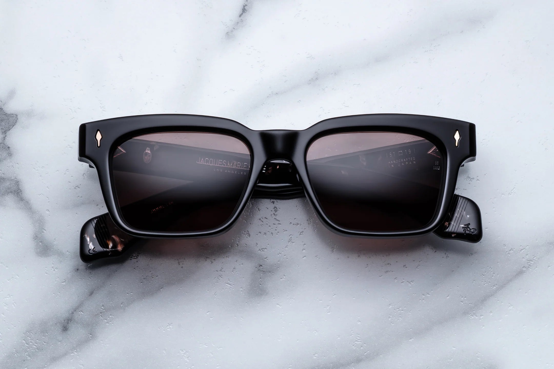 The Jacques Marie Mage Molino square sunglasses by Jacques Marie Mage feature a chunky, glossy black frame with dark lenses and small metallic accents at the corners. Placed on a white marble surface and viewed from the front, these bold designer glasses pay homage to Italian eyewear, offering a modern reinterpretation of classic rectangular shapes.