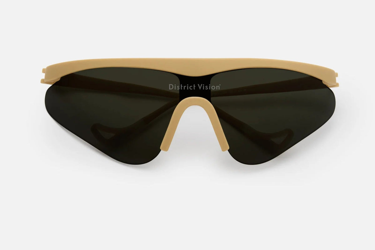 The District Vision Momoka Pacer sunglasses feature an ergonomic beige eyeglass frame paired with oversized, therapeutic-grade shatterproof polycarbonate lenses in a dark tint. The District Vision brand name is displayed on the lens, and the product is photographed against a plain white background.