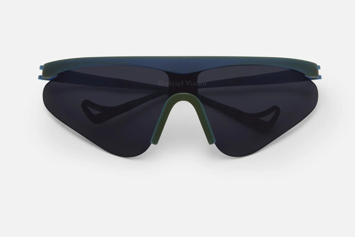 The District Vision Momoka Pacer by District Vision are dark, futuristic sports sunglasses featuring a single curved lens, a green nose bridge, and a minimal frame crafted from shatterproof polycarbonate, shown from the front against a plain white background.