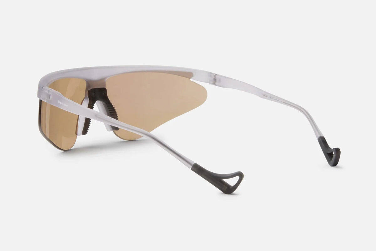 The District Vision Momoka Pacer by District Vision is a pair of futuristic, wraparound sunglasses featuring a lightweight TR90 frame and brown-tinted, therapeutic grade lenses, with thin silver arms, shown from a rear side angle on a white background.