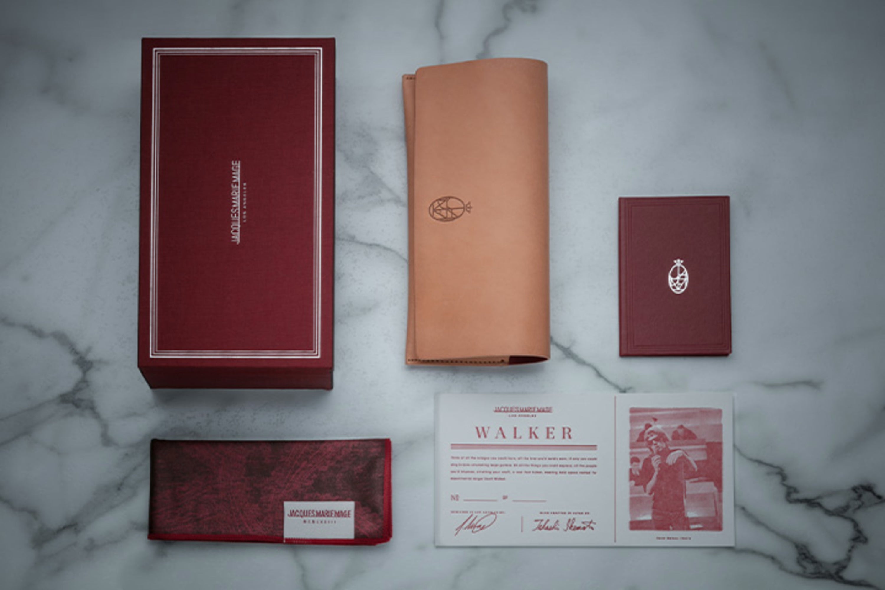 A Jacques Marie Mage Amilcar luxury eyewear box set is displayed on marble, featuring a red box, tan leather pouch, cardholder, cleaning cloth, certificate, and photo card—all meticulously arranged with Jacques Marie Mage branding in clear view. The presentation radiates sophisticated design and reflects the artisanal craftsmanship for which the brand is renowned.