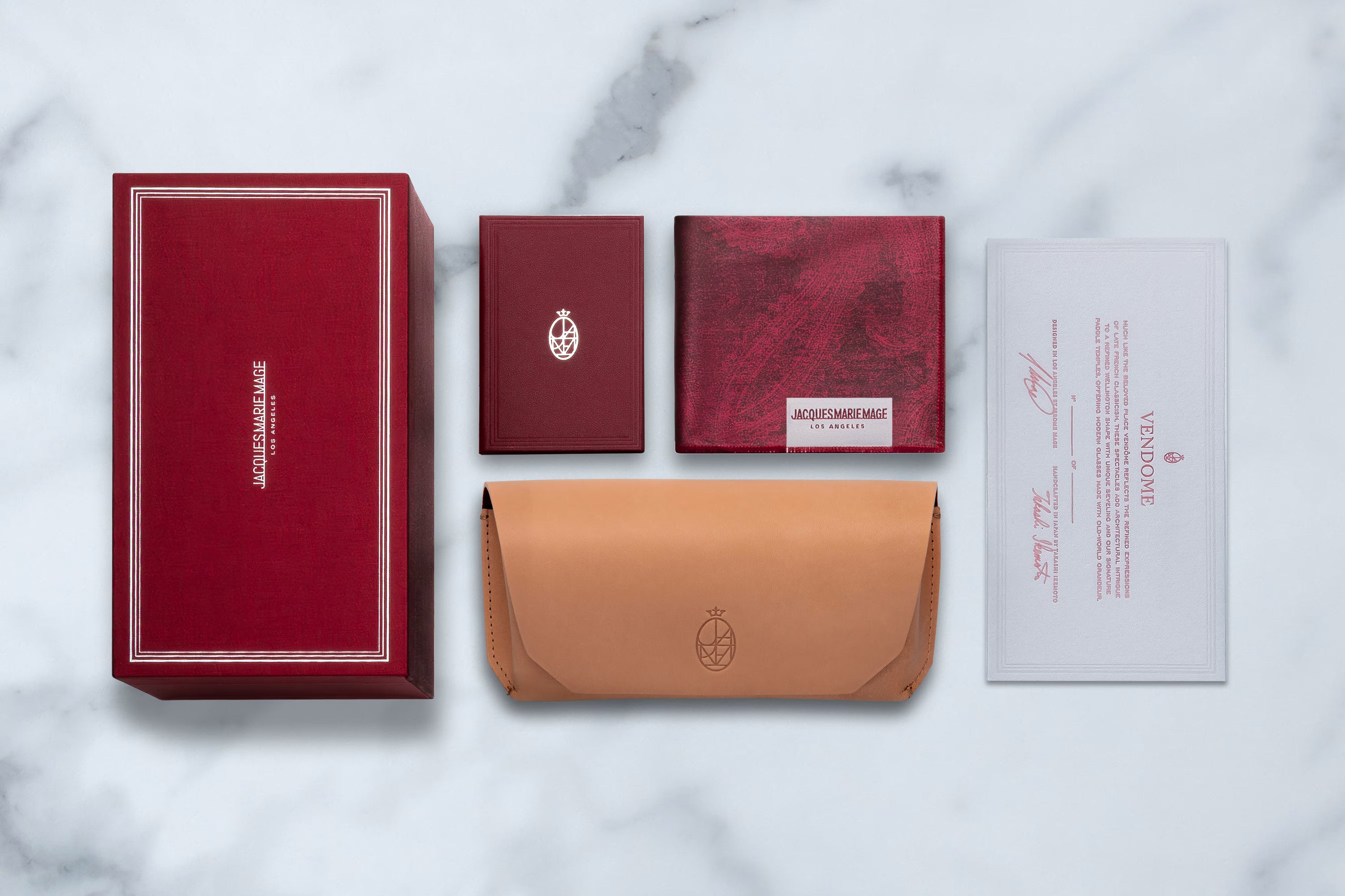 Arranged neatly on a marble surface are the Jacques Marie Mage Clyde accessories: a red box, two red booklets, a tan leather pouch, and a certificate. Each item features elegant Jacques Marie Mage branding, gold accents, and sophisticated wrap-arounds that embody the luxury and refinement of the brand.