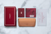 Arranged neatly on a marble surface are the Jacques Marie Mage Clyde accessories: a red box, two red booklets, a tan leather pouch, and a certificate. Each item features elegant Jacques Marie Mage branding, gold accents, and sophisticated wrap-arounds that embody the luxury and refinement of the brand.