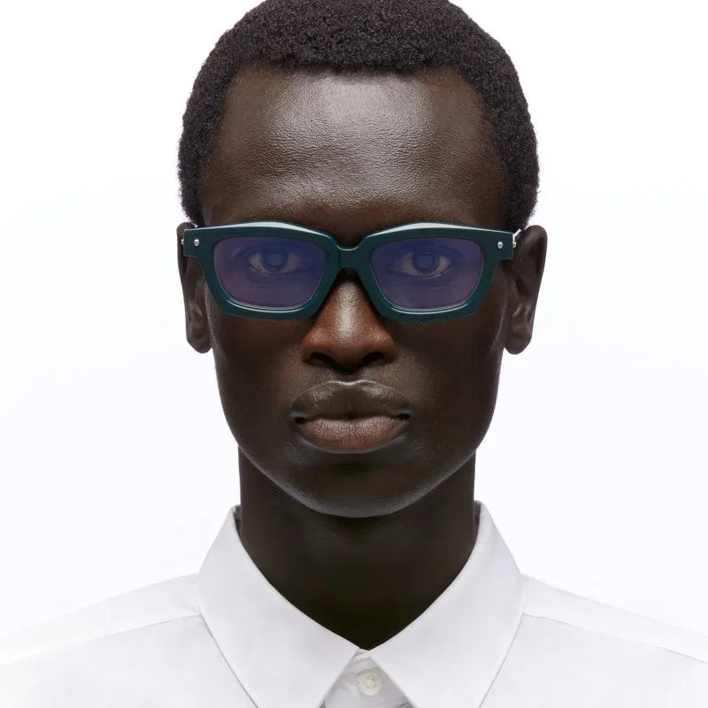 A close-up portrait of a man wearing thick, dark green square sunglasses with purple-tinted lenses and a white collared shirt against a plain white background.