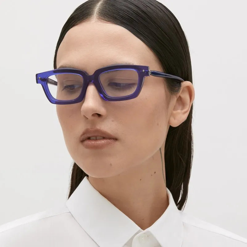 A person with straight dark hair, dressed in a white collared shirt and wearing bold blue rectangular Kuboraum P25 eyewear by Kuboraum, looks slightly downward against a plain light background.