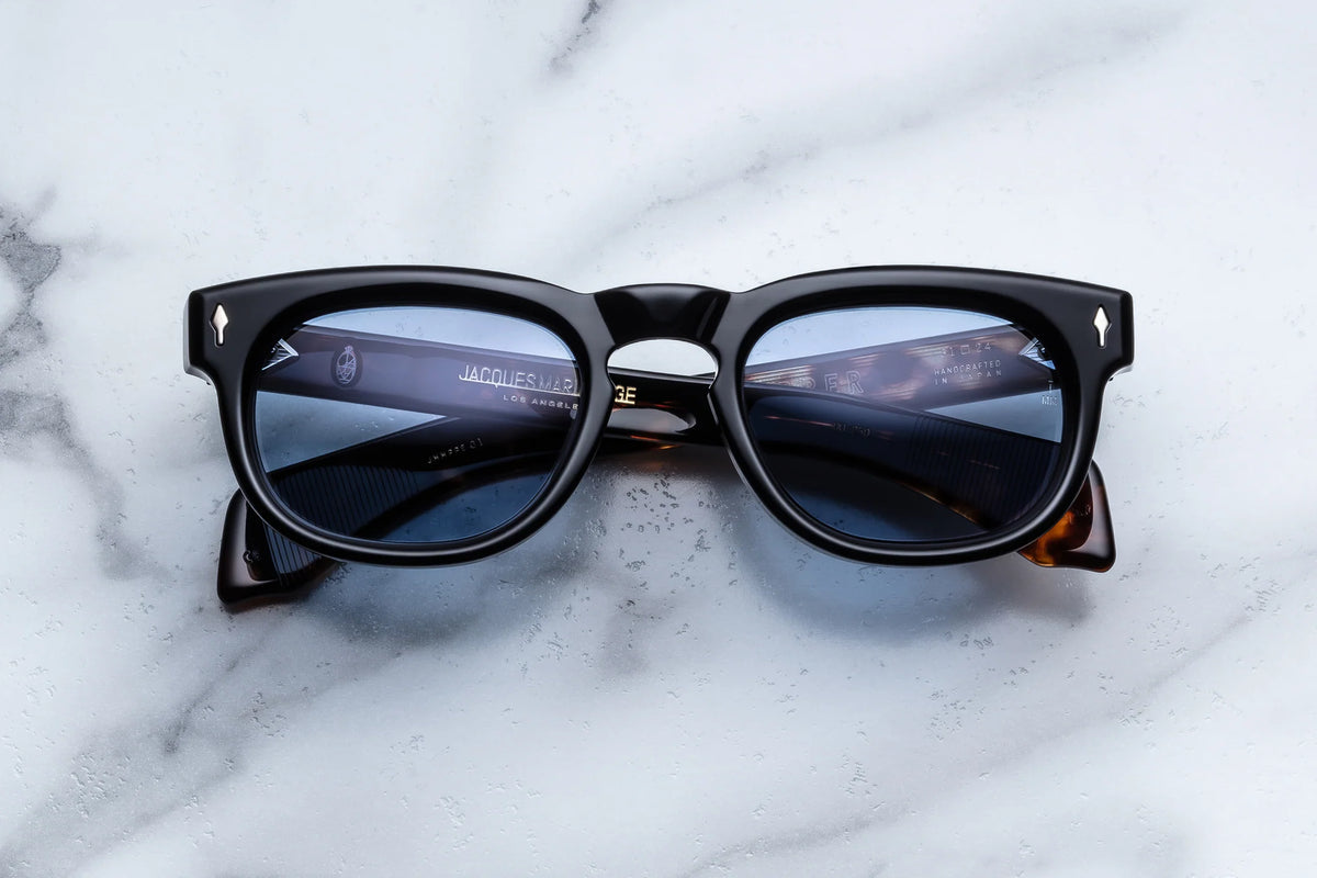 Black-rimmed Jacques Marie Mage A Decade of Hopper: Pepper sunglasses with tinted blue lenses, viewed from the front against a marble surface.