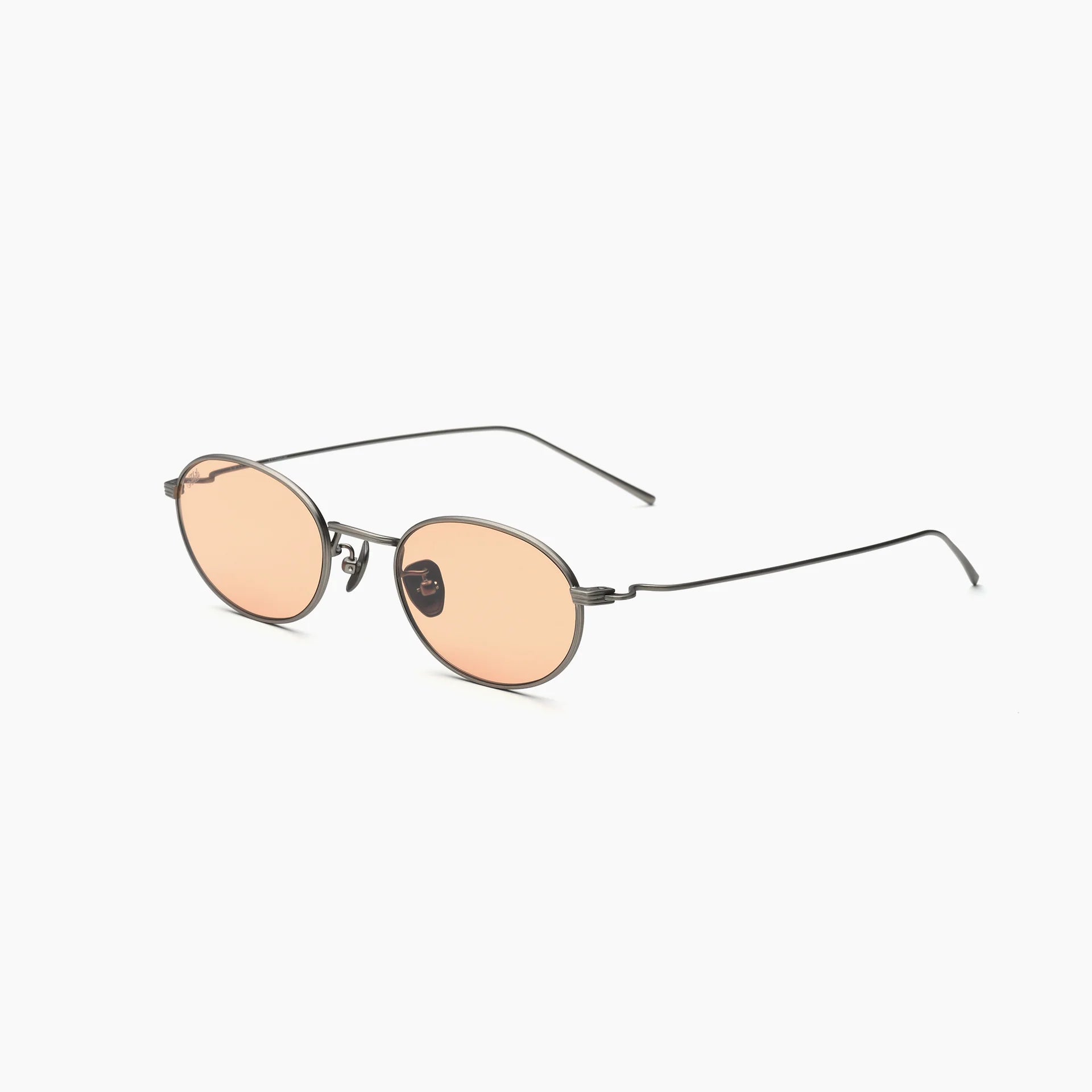Oval metal-frame sunglasses with peach-tinted lenses, adjustable nose pads, and slim wire temples, shown at an angled side view on a white background.