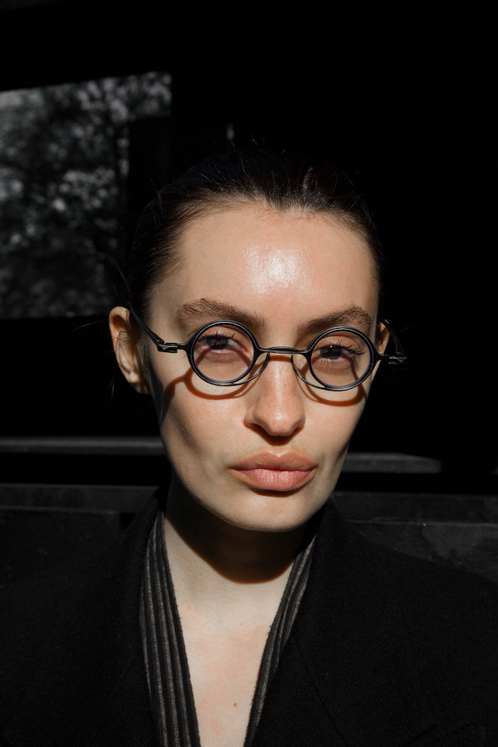Model wearing black RIGARDS round optical glasses with layered rims and a geometric bridge, photographed in dramatic sunlight.