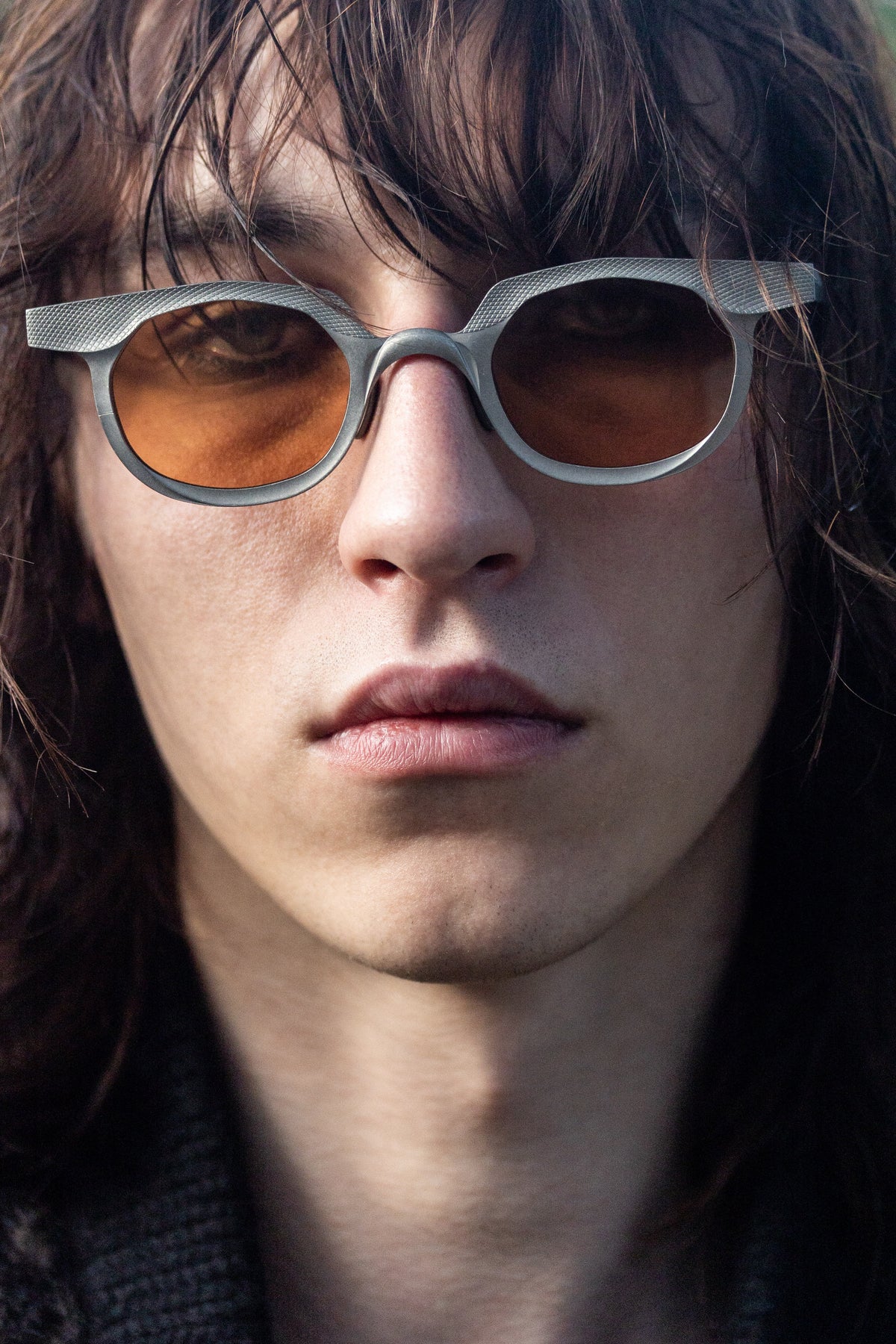 Model wearing silver RIGARDS sunglasses with orange lenses and textured brow detailing, photographed in a close-up portrait.