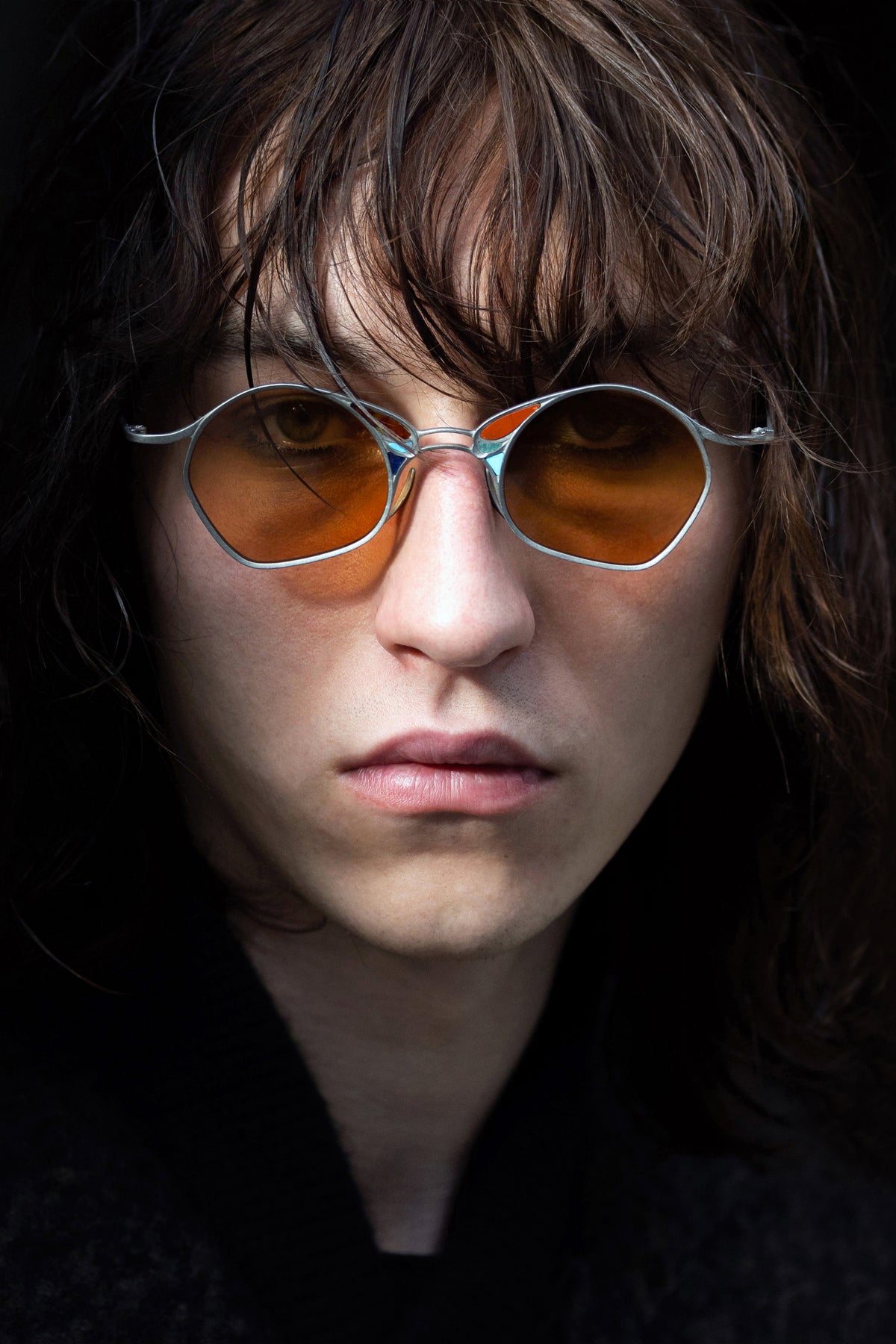 A person with long brown hair is wearing RIGARDS RG1032TI titanium sunglasses by RIGARDS, featuring round, orange-tinted lenses, and a black top, looking directly at the camera with a neutral expression.