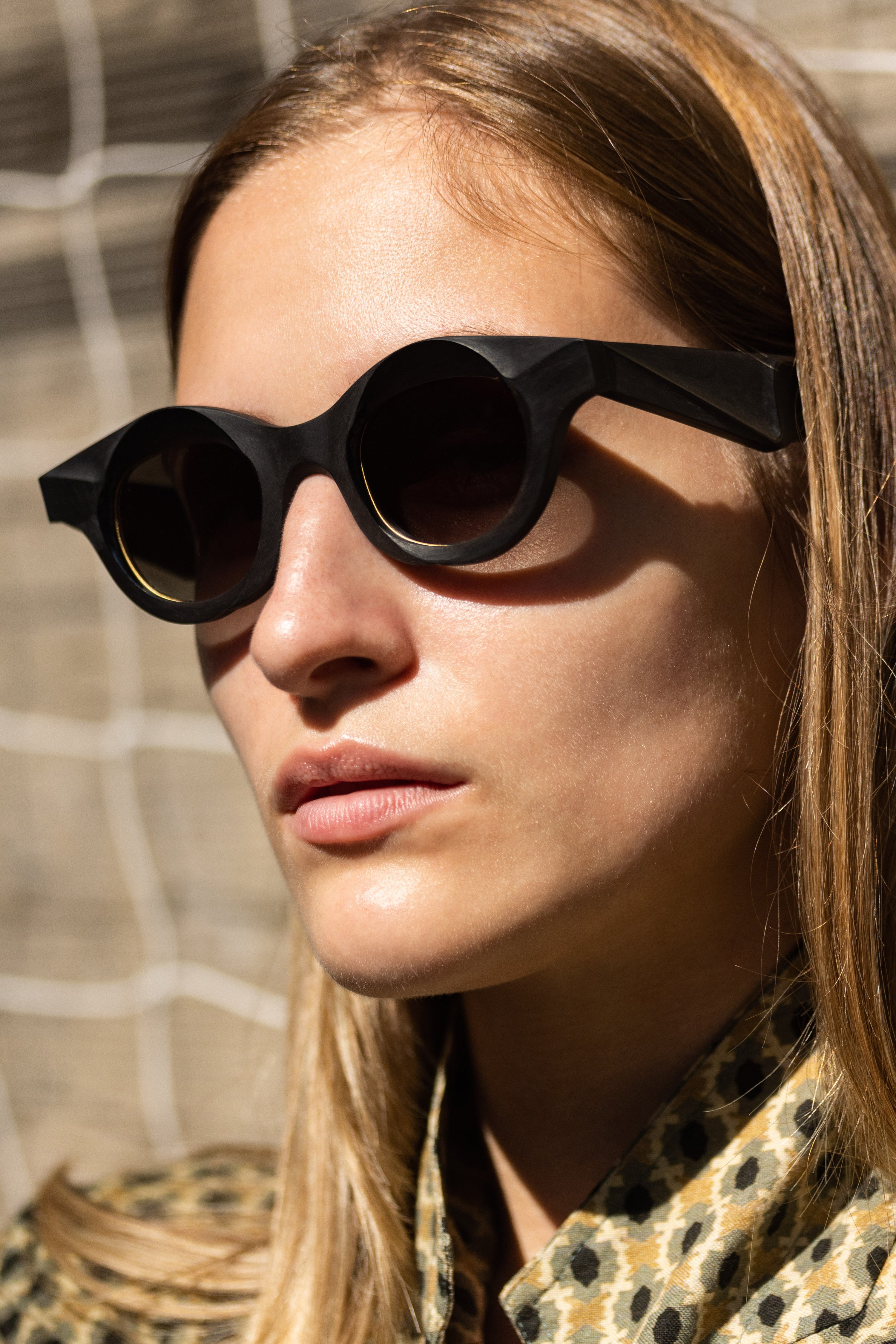 Model wearing sculptural black RIGARDS round sunglasses with dark lenses, photographed in sunlight from a three-quarter angle.