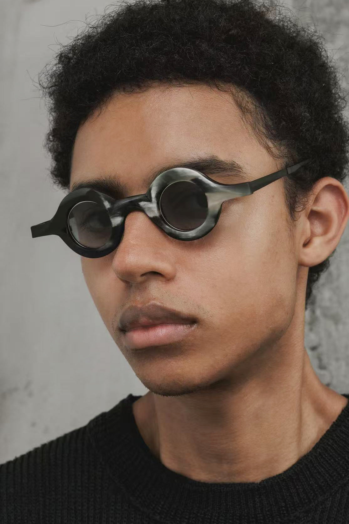 Model wearing sculptural black-and-grey horn RIGARDS sunglasses with round dark lenses, photographed from a three-quarter angle.
