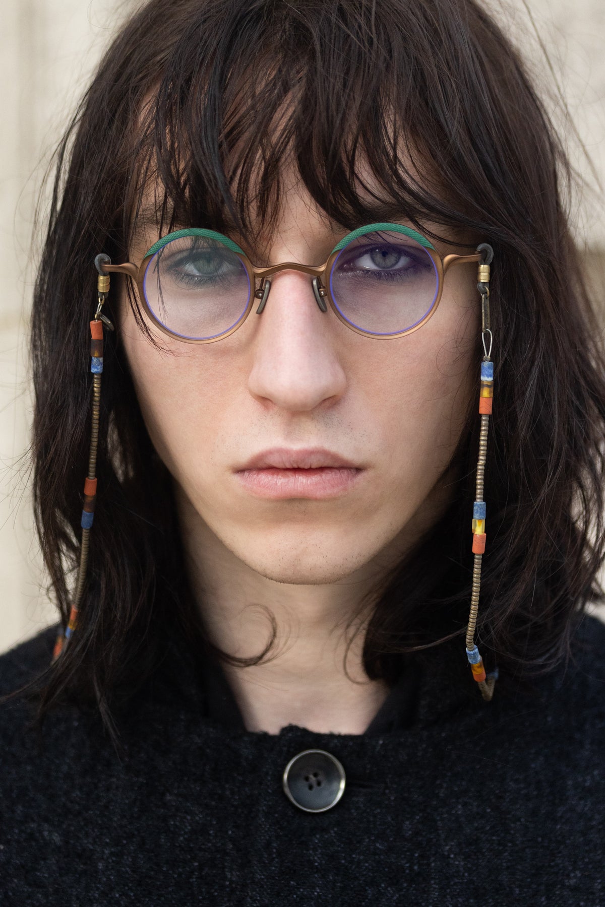 A person with long dark hair, wearing RIGARDS RG1199CU round glasses with UV400 sun lenses and beaded chains by RIGARDS, stares directly at the camera with a neutral expression. They are dressed in a dark, textured top featuring a single visible button.