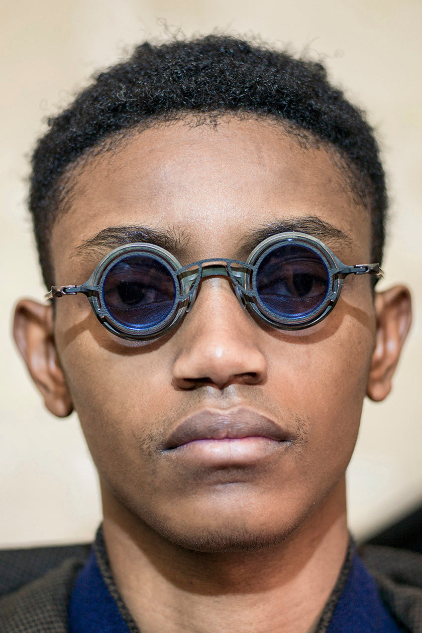 Model wearing blue RIGARDS round sunglasses with layered rims, dark lenses, and a geometric bridge, photographed in a close-up portrait.