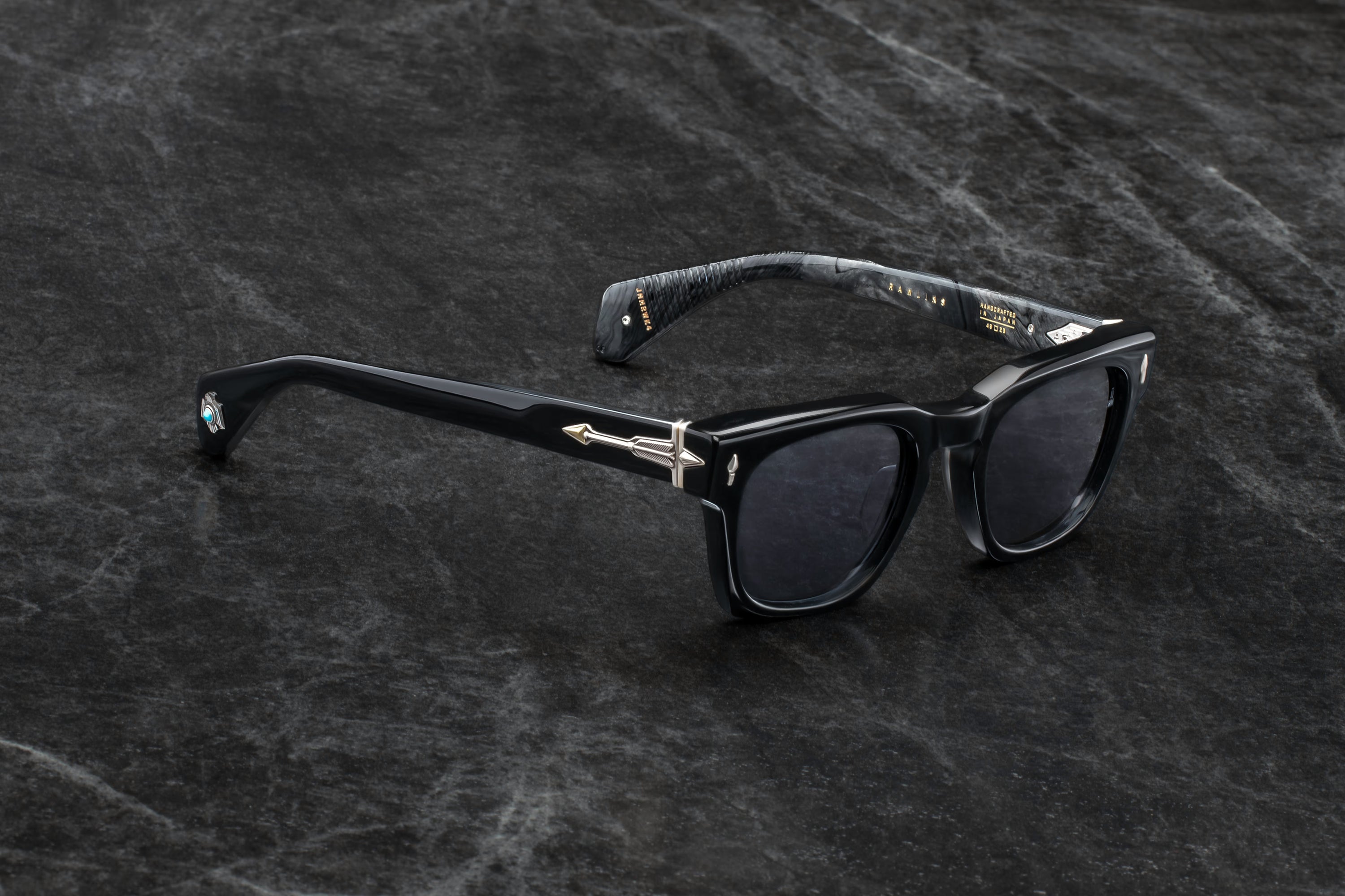 A pair of black acetate sunglasses with dark lenses and silver metal bridge details, resting on a dark textured stone surface.