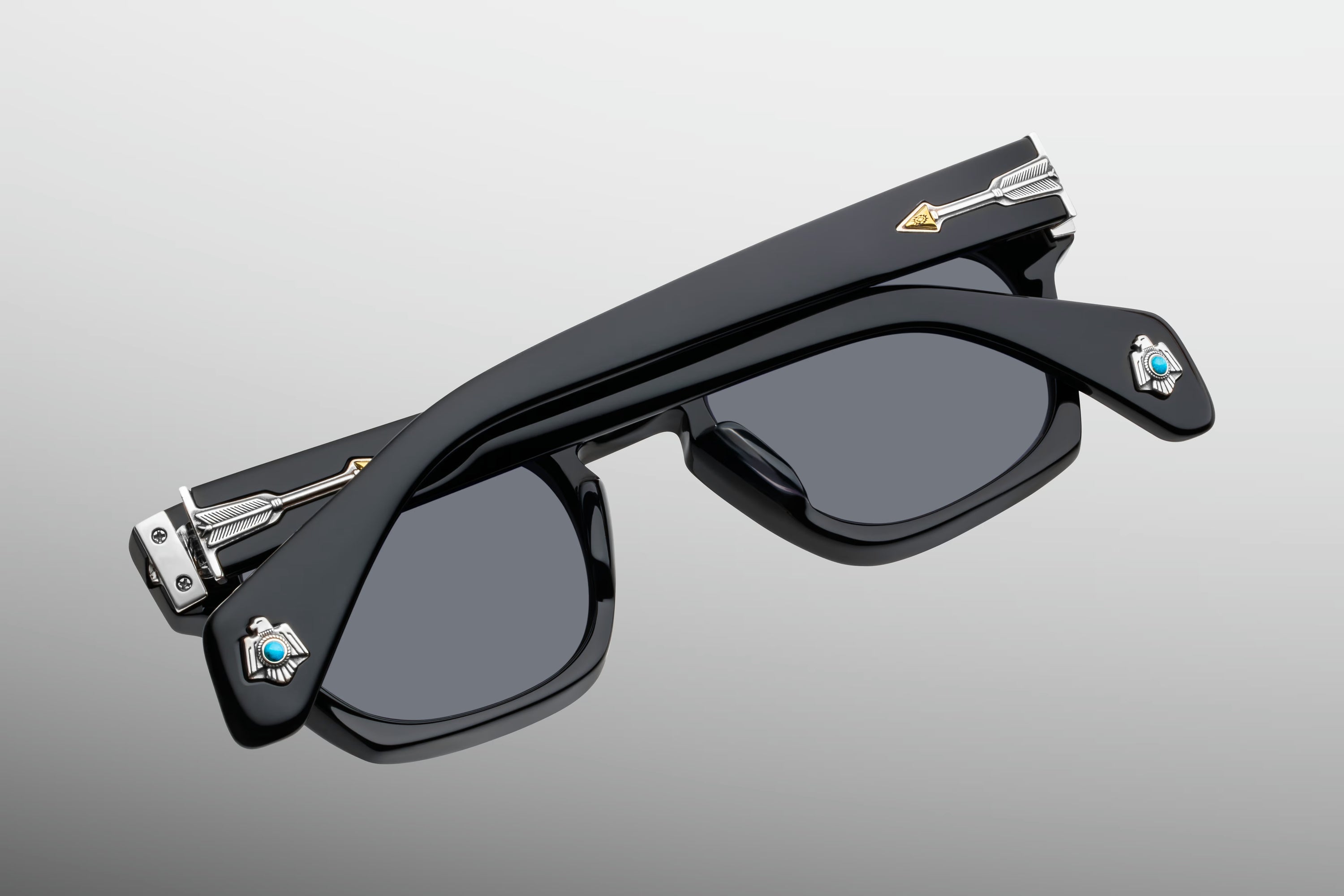 Black square sunglasses with dark lenses and small decorative blue and gold details on the temples, shown on a light gray gradient background.