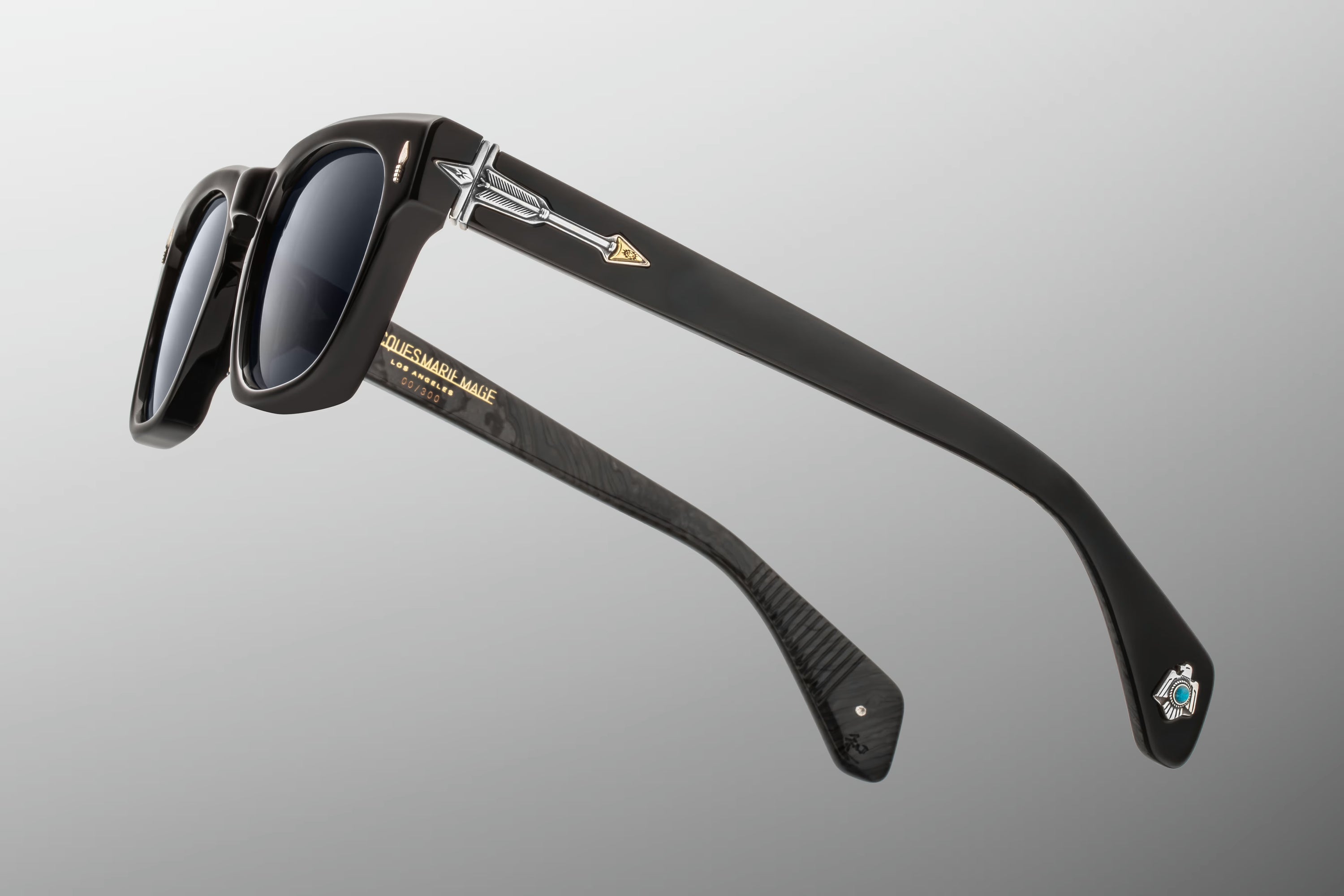 Black acetate sunglasses with dark lenses, featuring a gold arrow logo on the temple and a small blue and white emblem near the tip of the other arm, shown against a neutral gray background.
