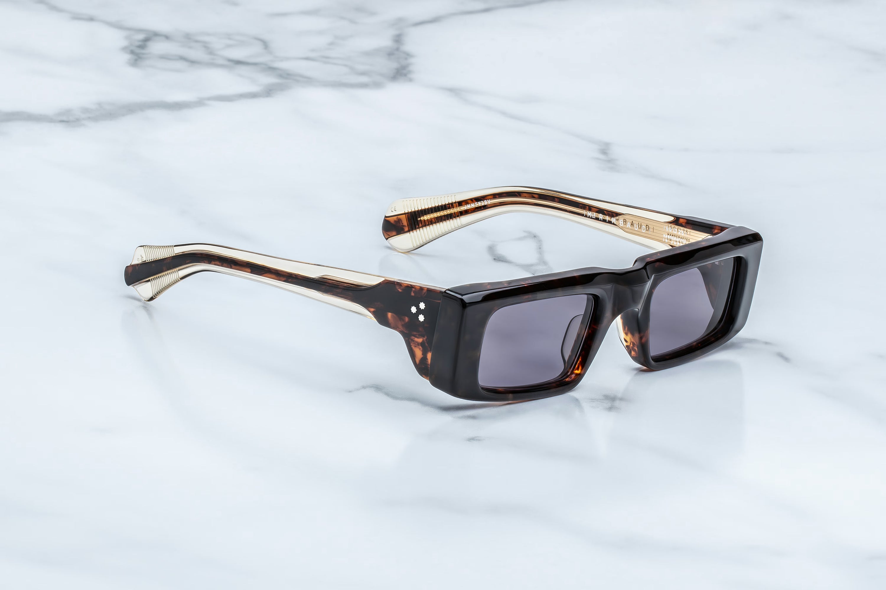The Jacques Marie Mage Rimbaud sunglasses by Jacques Marie Mage, featuring a tortoiseshell square frame and dark lenses evocative of the indie sleaze scene, rest on a white marble surface.