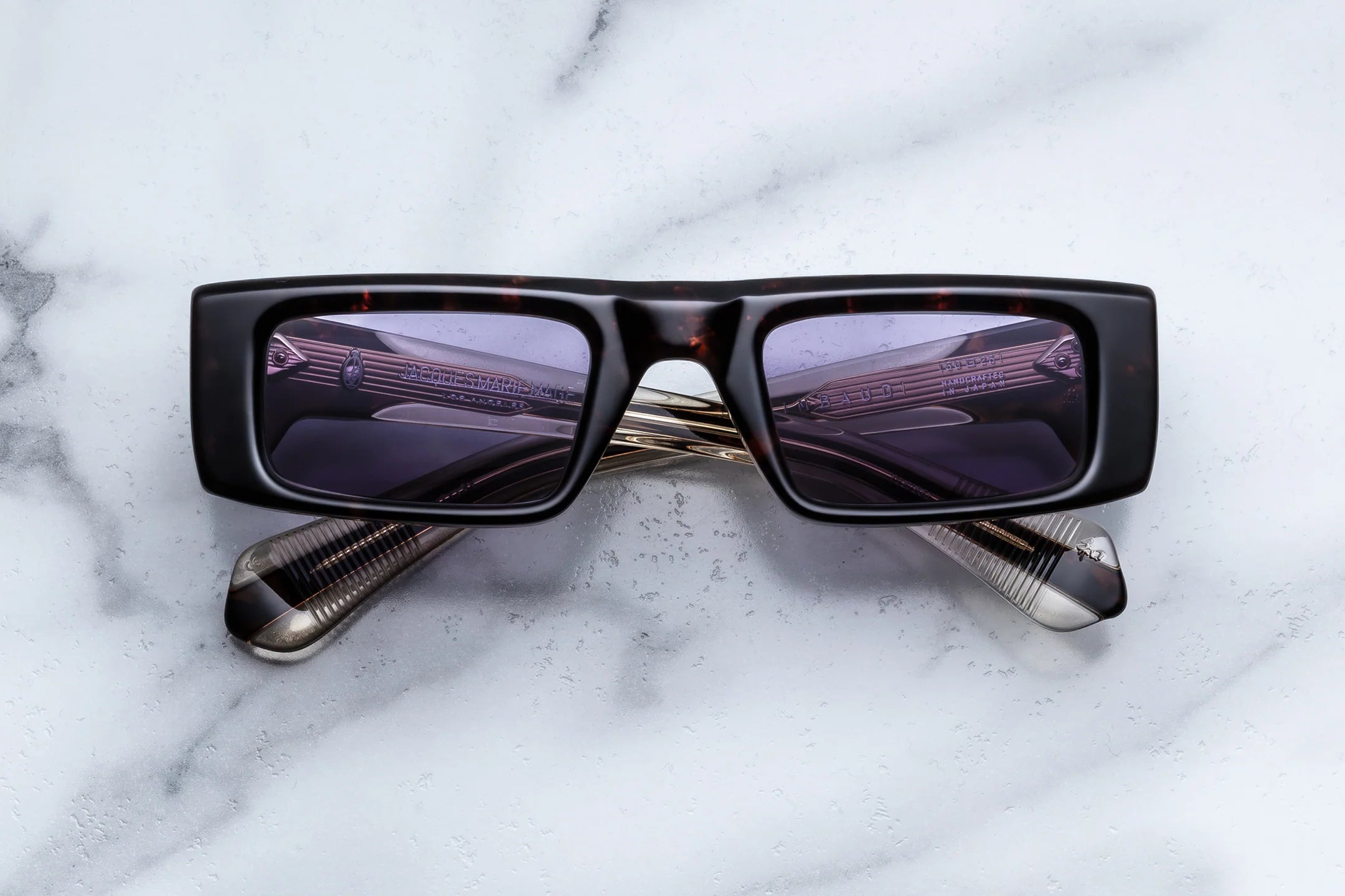 A pair of Jacques Marie Mage Rimbaud rectangular black sunglasses with dark lenses—evoking indie sleaze style—is displayed face up on a white marble surface, with the limited-edition arms crossed behind the lenses.