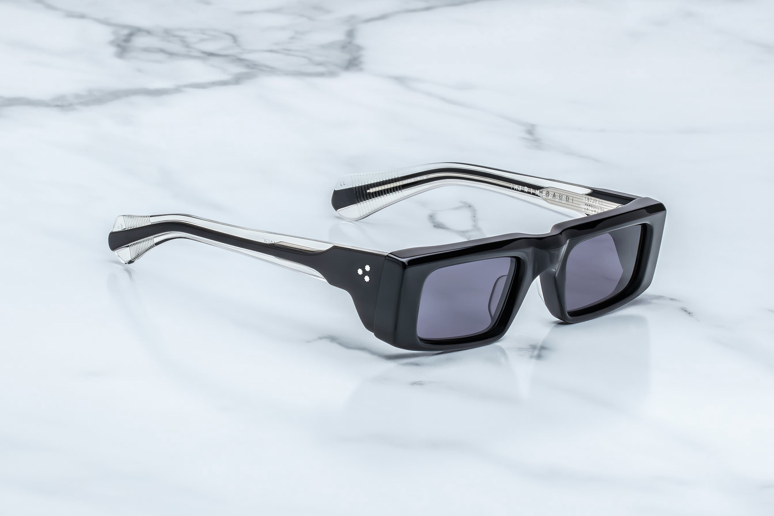 The Jacques Marie Mage Rimbaud sunglasses, featuring black rectangular frames with bold, thick edges and dark lenses, are displayed on a white marble surface veined with subtle grey—these limited-edition spectacles from Jacques Marie Mage evoke the cool edge of garage rock revival style.