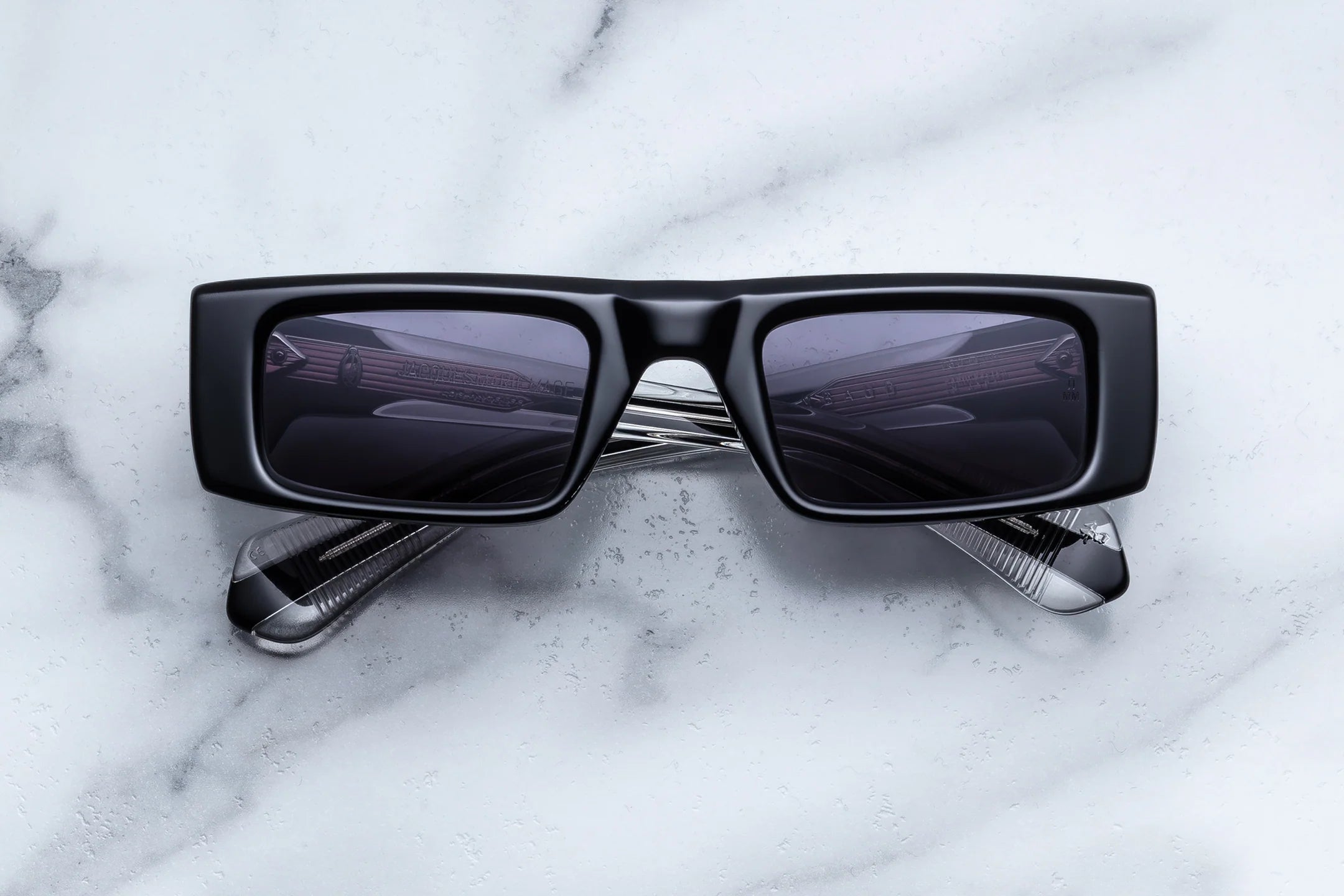 A pair of Jacques Marie Mage Rimbaud black rectangular sunglasses by Jacques Marie Mage, evocative of limited-edition eyewear, is displayed atop two crossed golf clubs on a white marble surface.