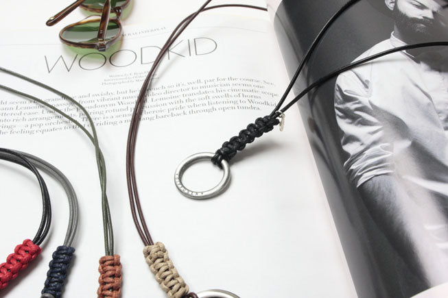 A close-up flat lay of several corduroy necklaces with silver-toned circular pendants, arranged on an open magazine. One necklace has a dark brown cord, one has a black cord, and others in red, navy, and tan are partially visible.