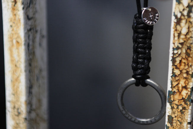 A close-up of a black paracord-wrapped keychain or bag charm with a silver metal ring and a round silver tag, hanging against a dark gray background.