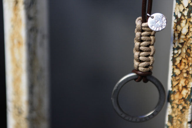 A close-up of a paracord-wrapped keychain or bag charm with a silver metal ring and a small round silver tag, hanging against a dark gray background.