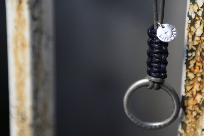 A close-up of a dark navy paracord-wrapped metal ring accessory with a small silver coin charm, hanging against a dark blurred background.
