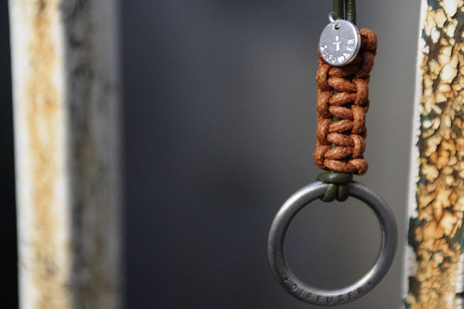 A close-up of a brown paracord-wrapped loop accessory with a silver metal ring and a small round silver charm, shown against a dark blurred background.