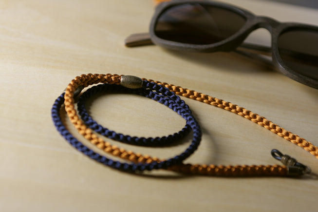 A braided sunglasses strap in navy blue and tan, displayed on a light wood surface next to a pair of sunglasses.
