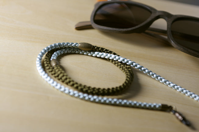 A close-up of a braided chain strap in silver and bronze tones, coiled on a light wood surface next to a pair of dark sunglasses.