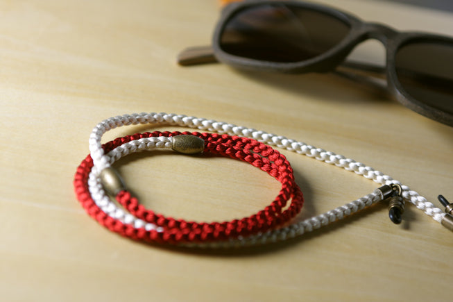 A braided rope neck strap or lanyard in red and white with small metallic silver and gold-toned beads, displayed on a light wood surface next to a pair of sunglasses.