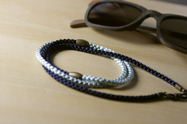 A braided eyeglasses strap in navy blue and silver with brass-toned metal accents, displayed on a light wood surface next to sunglasses.