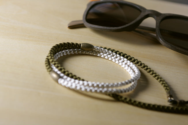 A close-up of a multi-strand braided leather strap accessory in olive green and silver, resting on a light wood surface next to a pair of sunglasses.