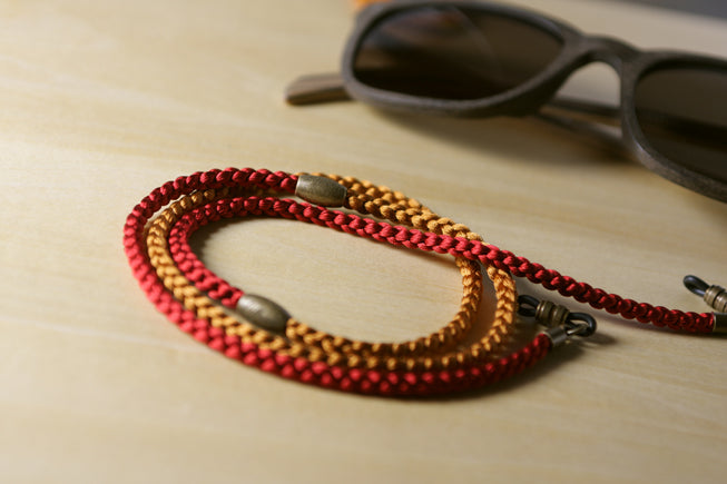 A braided red and gold eyeglass strap with metal hardware, displayed on a light wood surface next to a pair of sunglasses.