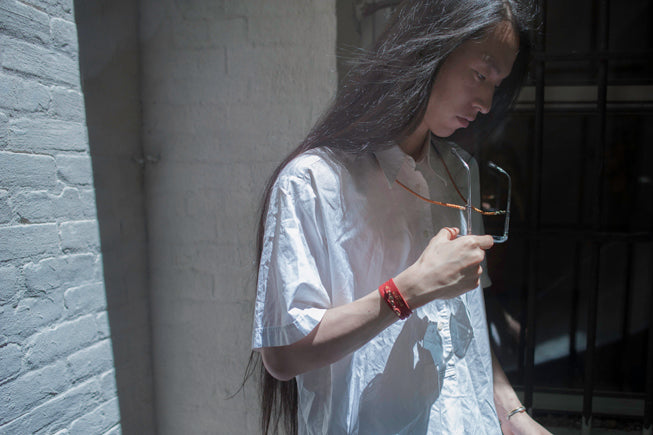 A person with long dark hair wearing a loose white short-sleeve shirt, holding a pair of eyeglasses in a dimly lit interior next to a white brick wall.