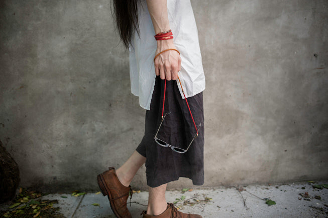 A person wearing a white t-shirt, a dark gray midi skirt, and brown lace-up boots walks against a textured gray concrete wall while carrying a small black shoulder bag with red straps.