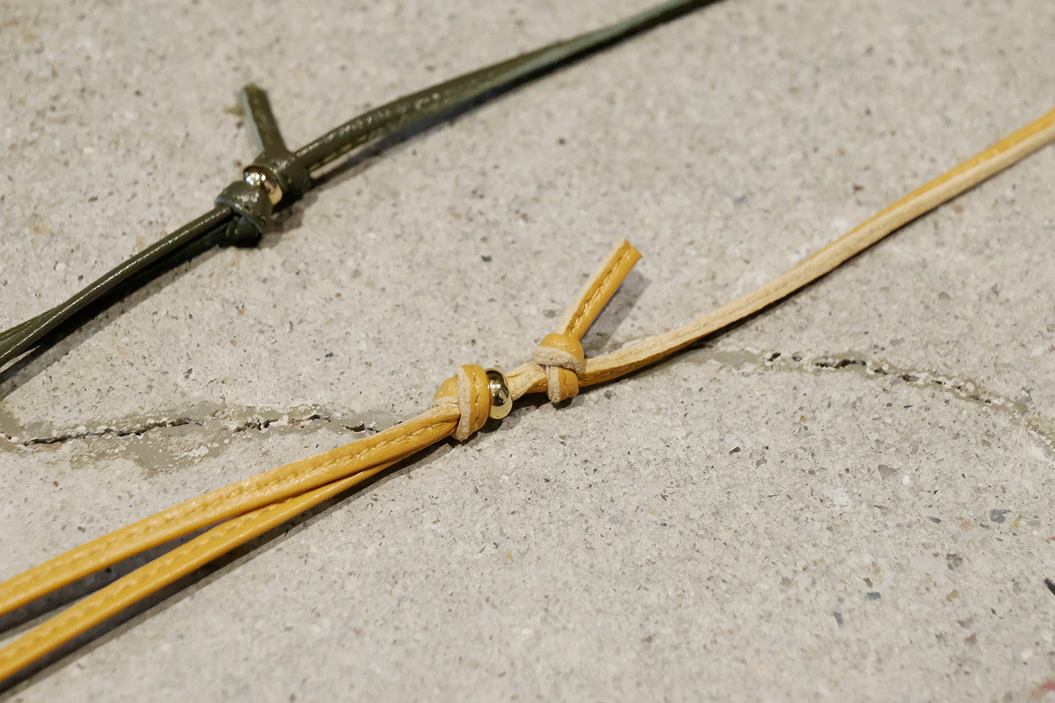 A close-up of a handmade leather cord or belt tie with a knot, laid on a textured gray concrete surface next to a dark metal rod.